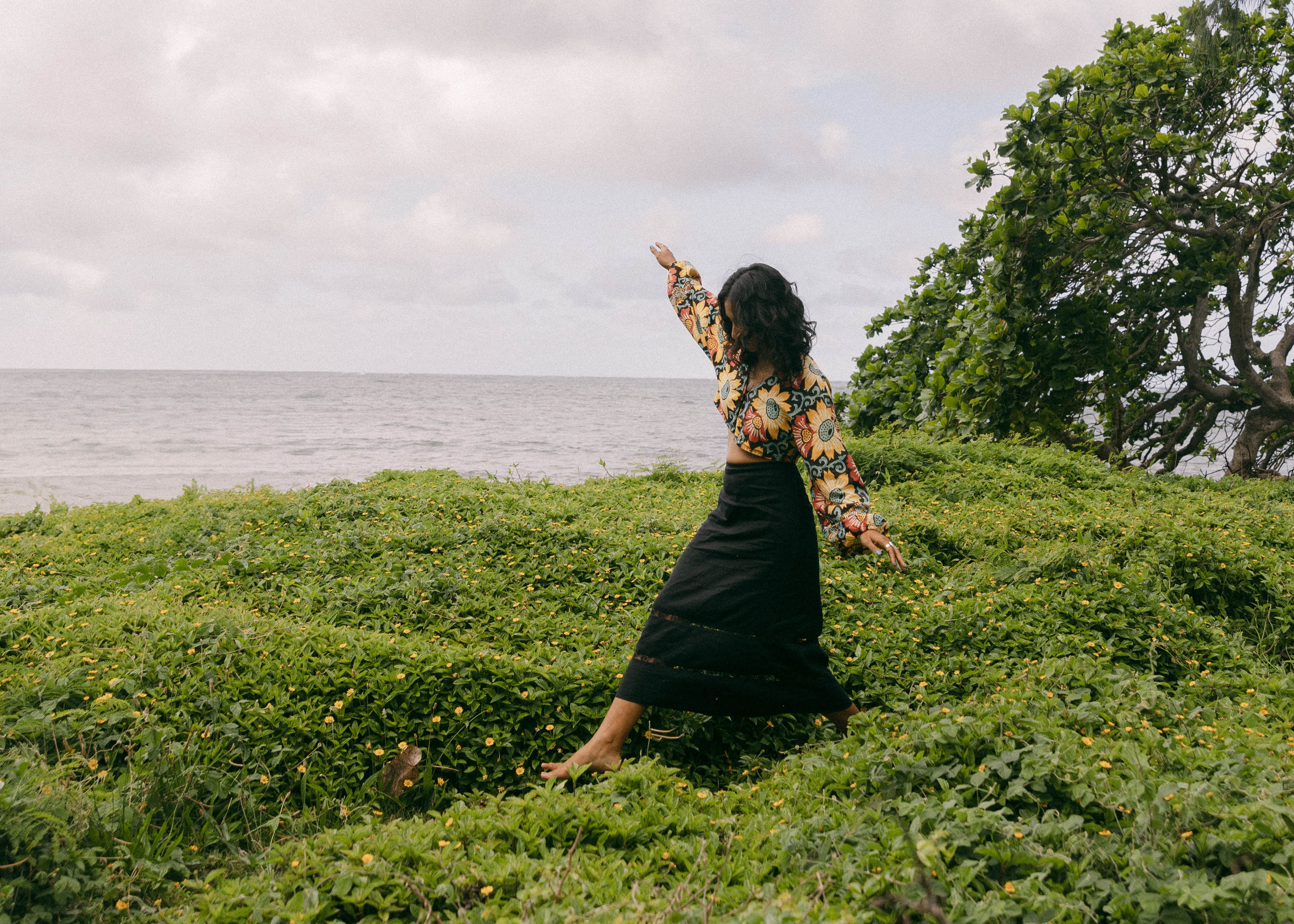 A woman in a black dress and patterned top poses on a grassy hill overlooking the ocean under a cloudy sky.