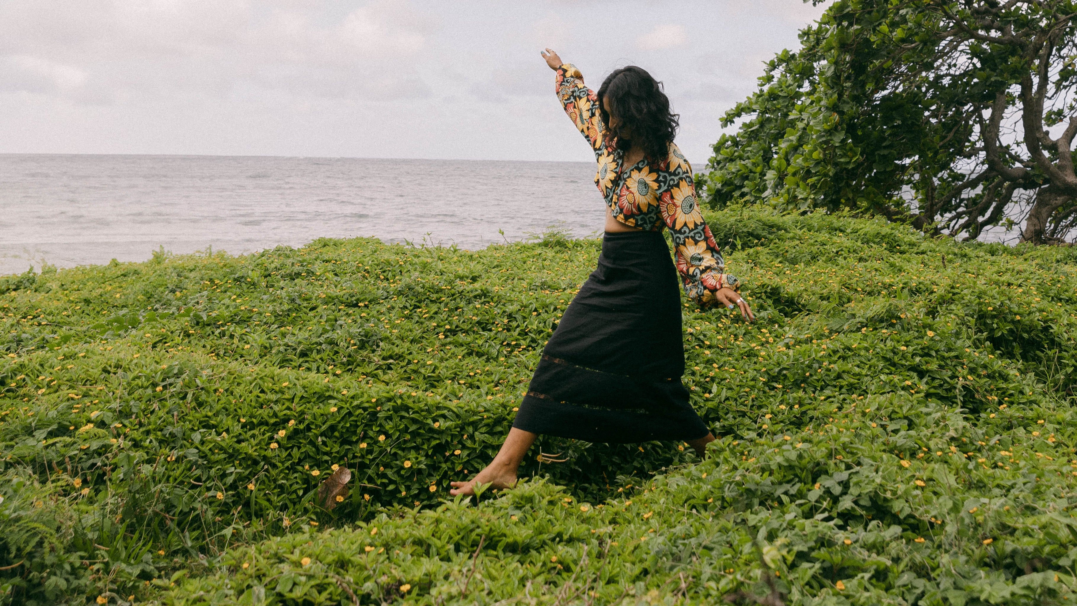 A woman in a black dress and patterned top poses on a grassy hill overlooking the ocean under a cloudy sky.