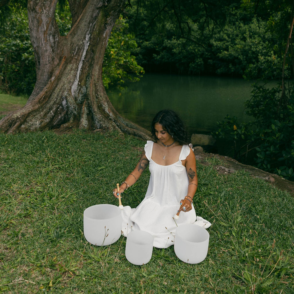 A woman in a white sleeveless dress sits on grass outdoors by a lake, holding three white stone singing bowls with wooden sticks. A large tree trunk is visible behind her, with dense green foliage and water in the background.