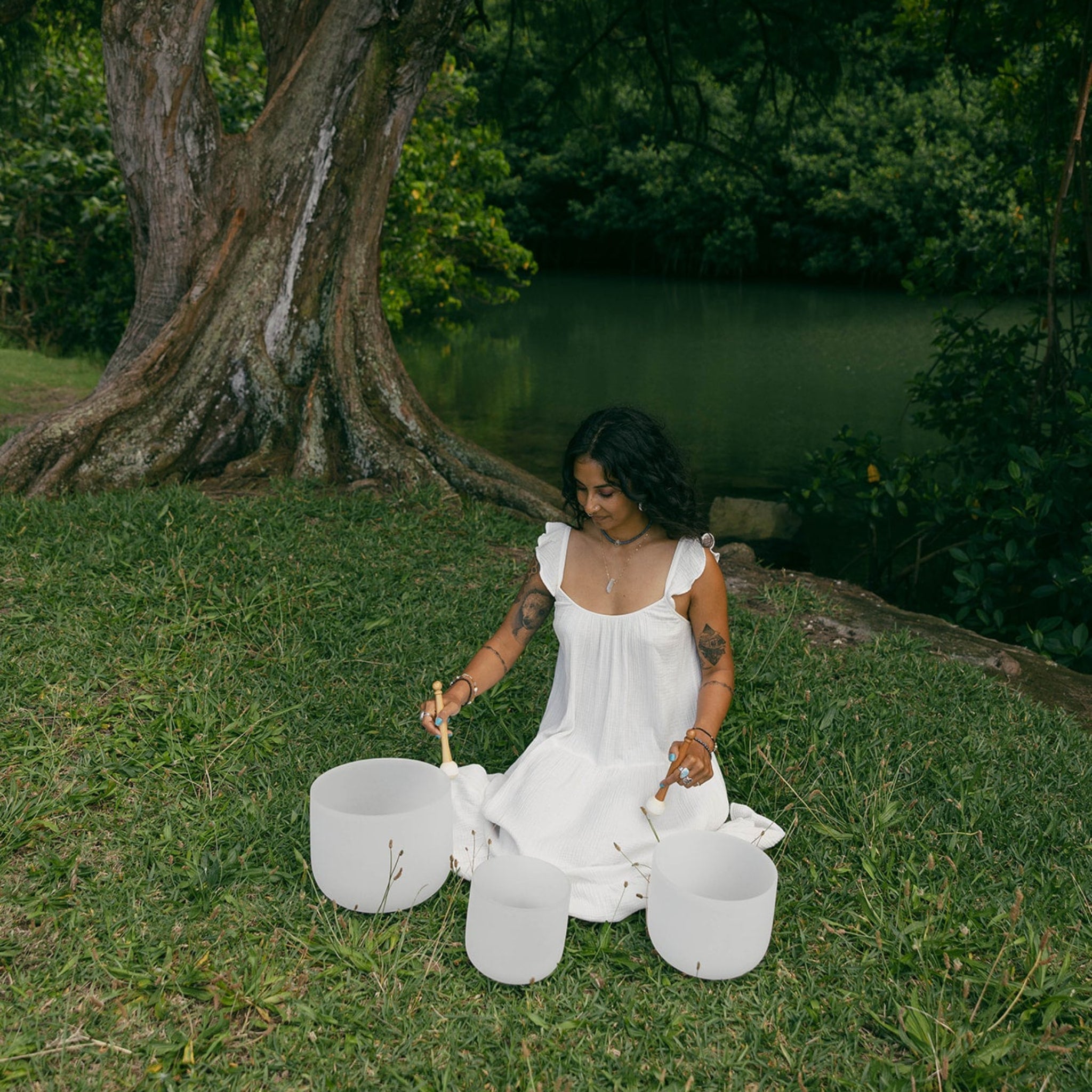 A woman in a white sleeveless dress sits on grass outdoors by a lake, holding three white stone singing bowls with wooden sticks. A large tree trunk is visible behind her, with dense green foliage and water in the background.
