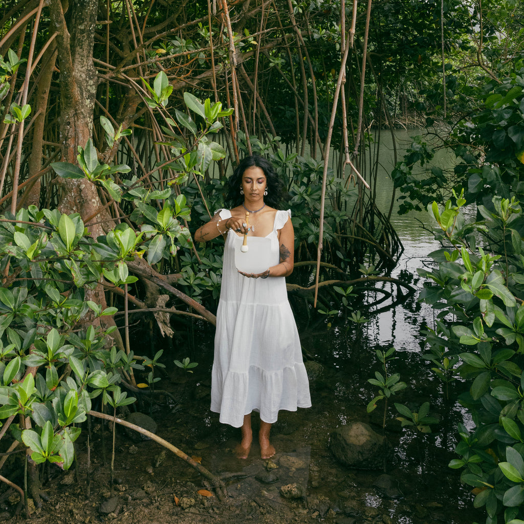 A woman wearing a white sleeveless tiered maxi dress and a gold pendant necklace stands barefoot in a muddy, water-filled mangrove swamp surrounded by dense green foliage.