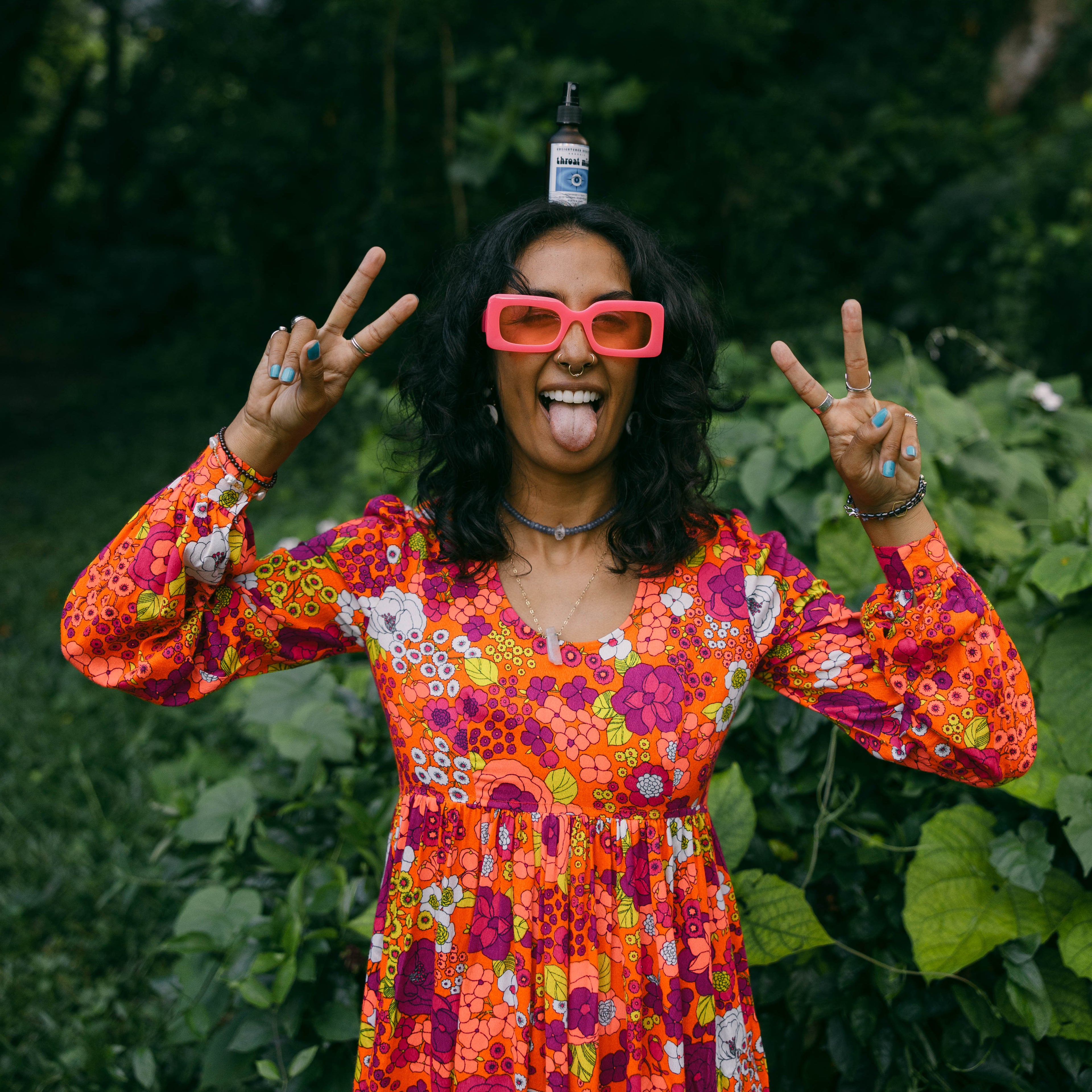 A woman outdoors in a lush green setting, wearing a bright orange and red floral long-sleeve dress, pink novelty sunglasses, and balancing a small bottle on her head while making peace signs with both hands.