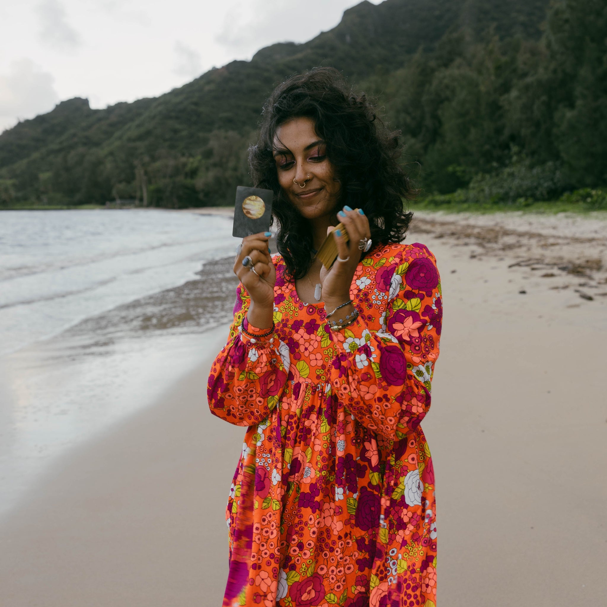 A woman stands on a sandy beach with water and a forested mountain in the background, holding up a small square jewelry piece to inspect it. She is wearing a long-sleeve, vibrant floral dress with orange, pink, purple, and yellow tones.