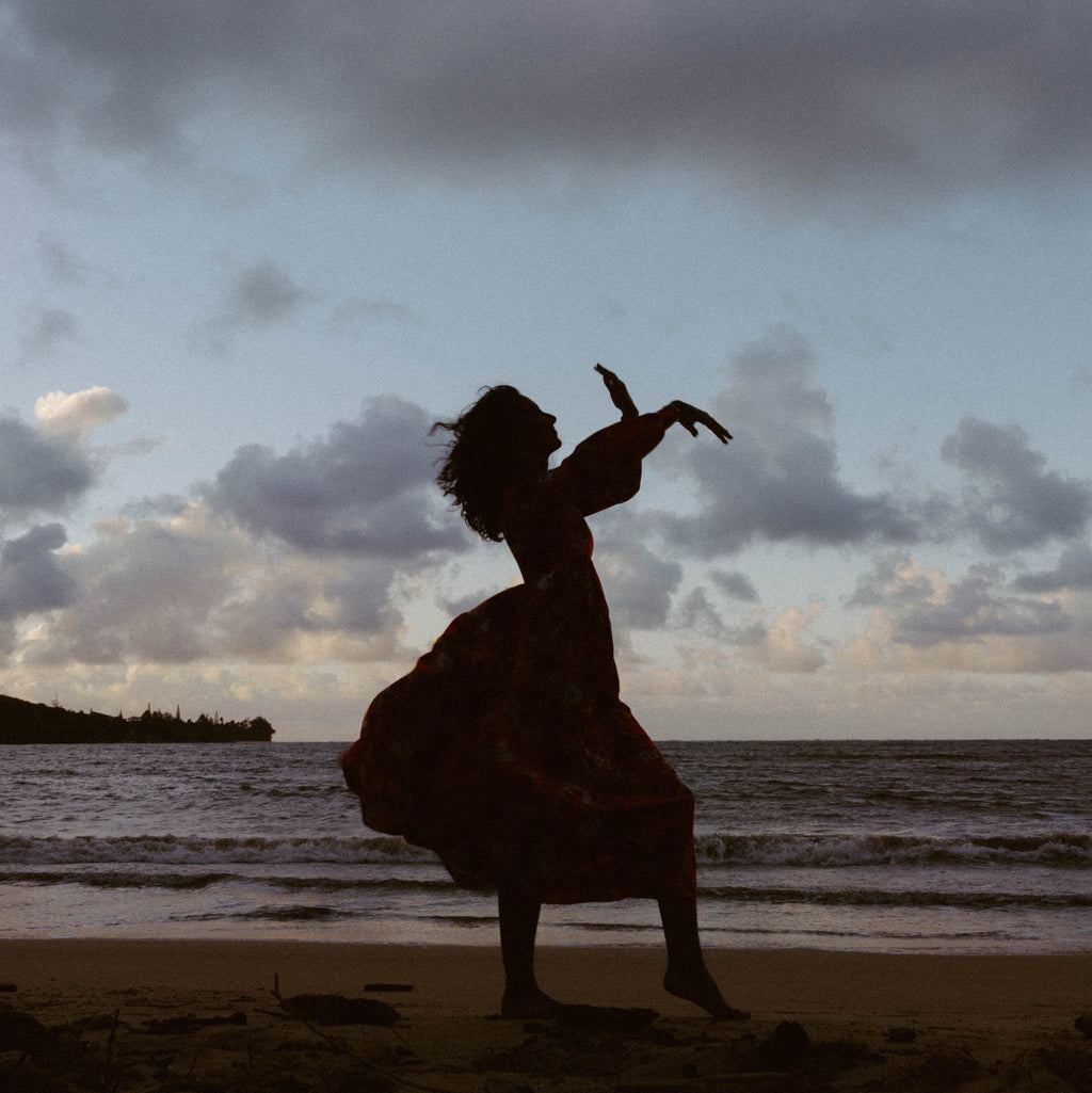 A woman in a flowing dress stands in silhouette on a beach, gesturing with her hands against a cloudy sky and ocean backdrop.