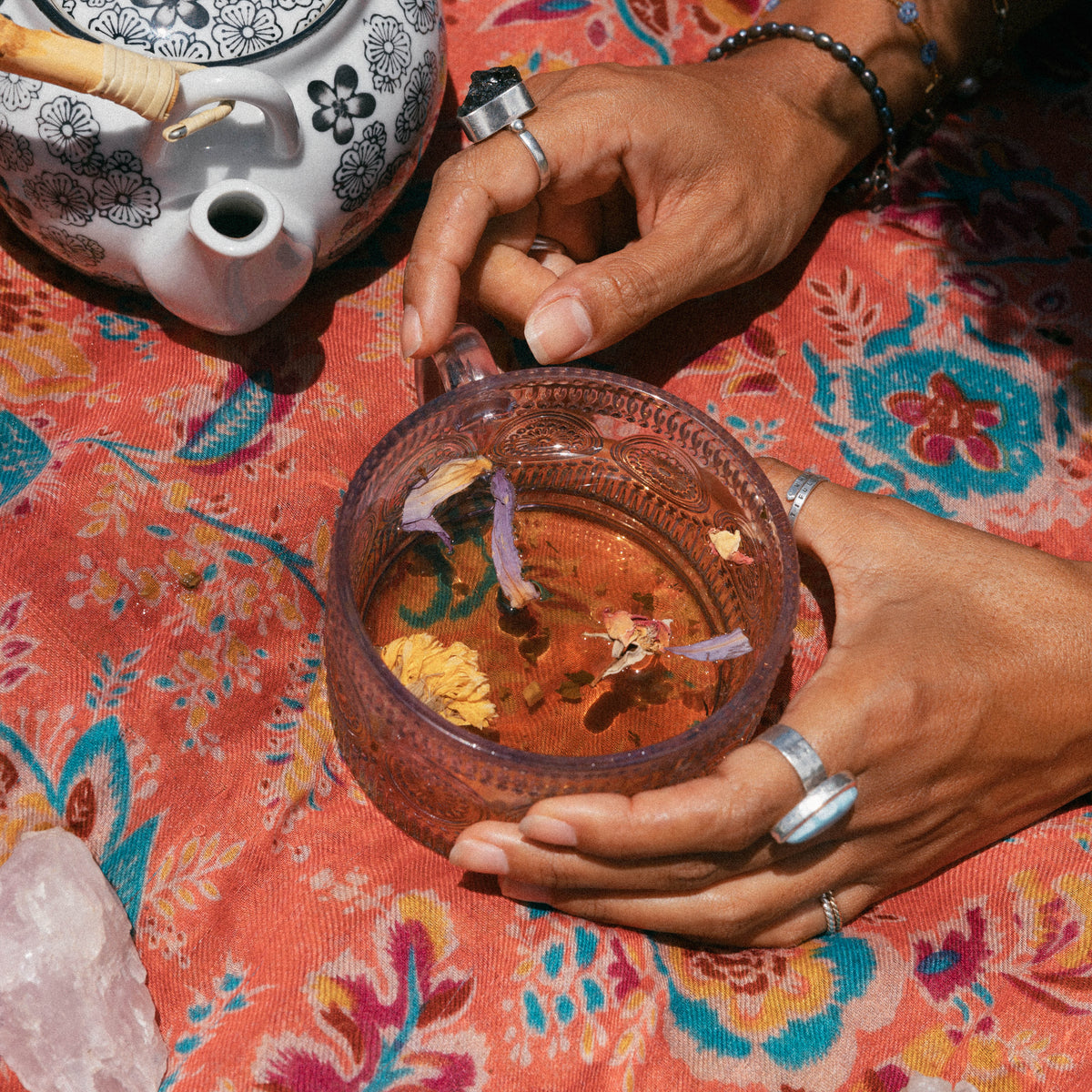 A close-up of hands holding a translucent amber glass mug filled with herbal tea and flower petals, set on a vibrant orange and pink floral blanket next to a white patterned teapot.