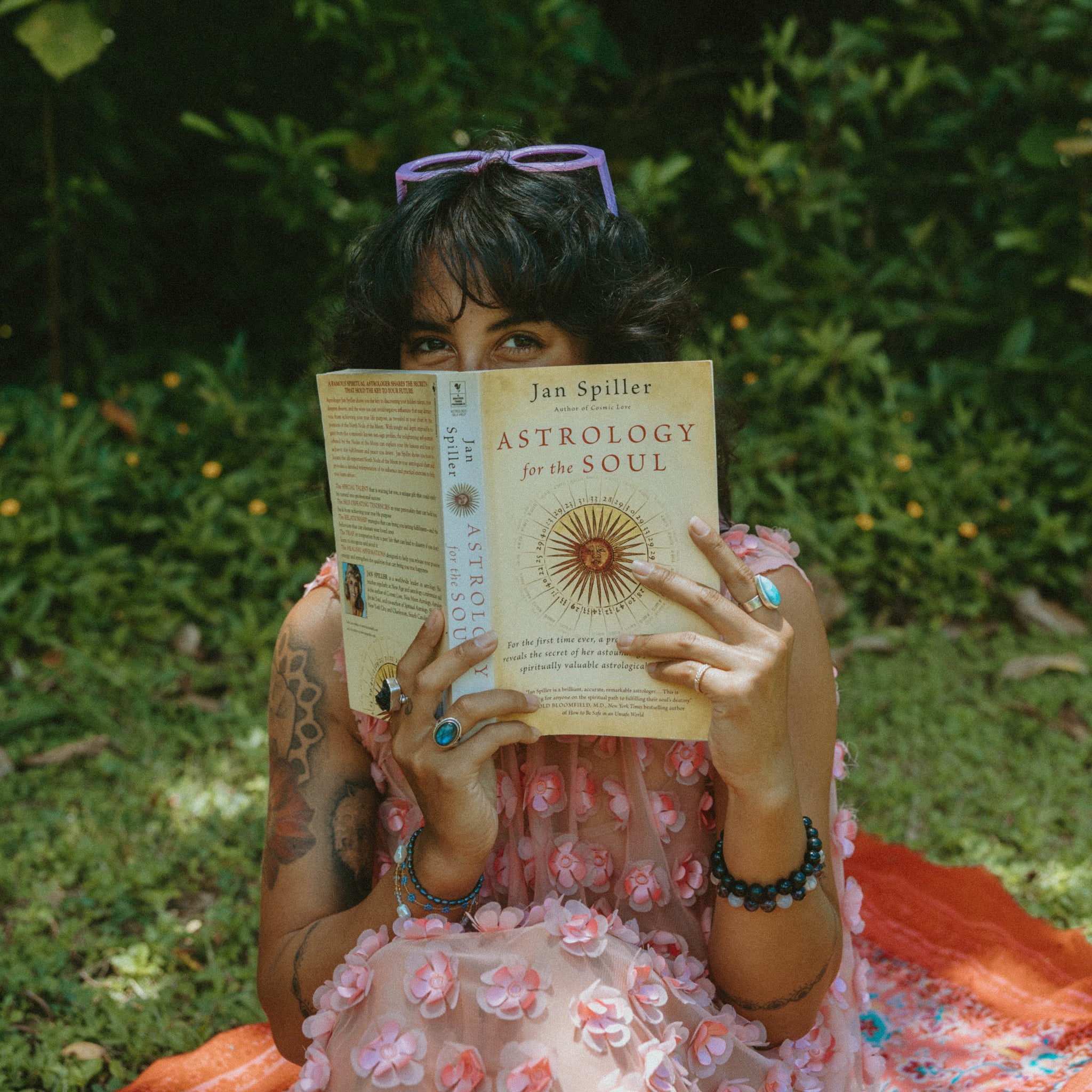 A woman sitting outdoors on a red blanket in a grassy, leafy area, holding a book titled 'ASTROLOGY FOR THE SOUL' by Jan Spiller. She is wearing a pink floral dress and has purple sunglasses on her head.