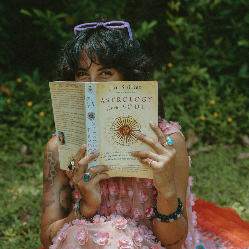 A woman sitting outdoors in a garden setting, holding a copy of the book 'Astrology for the Soul' by Jan Spiller. She is wearing a pink floral dress and has purple sunglasses on her head.