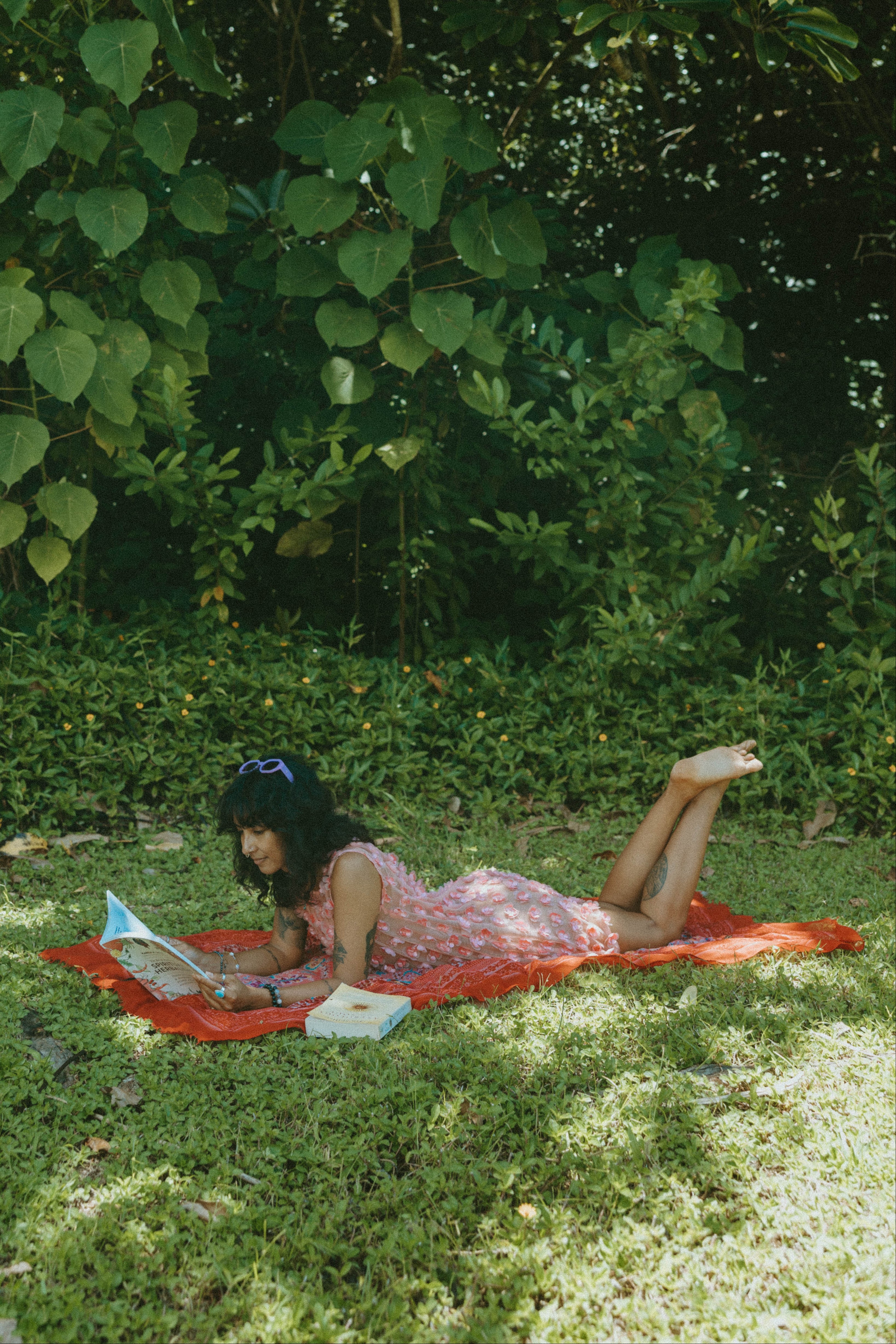 A woman in a pink floral dress lies on a red blanket reading on a grassy lawn surrounded by lush green trees.