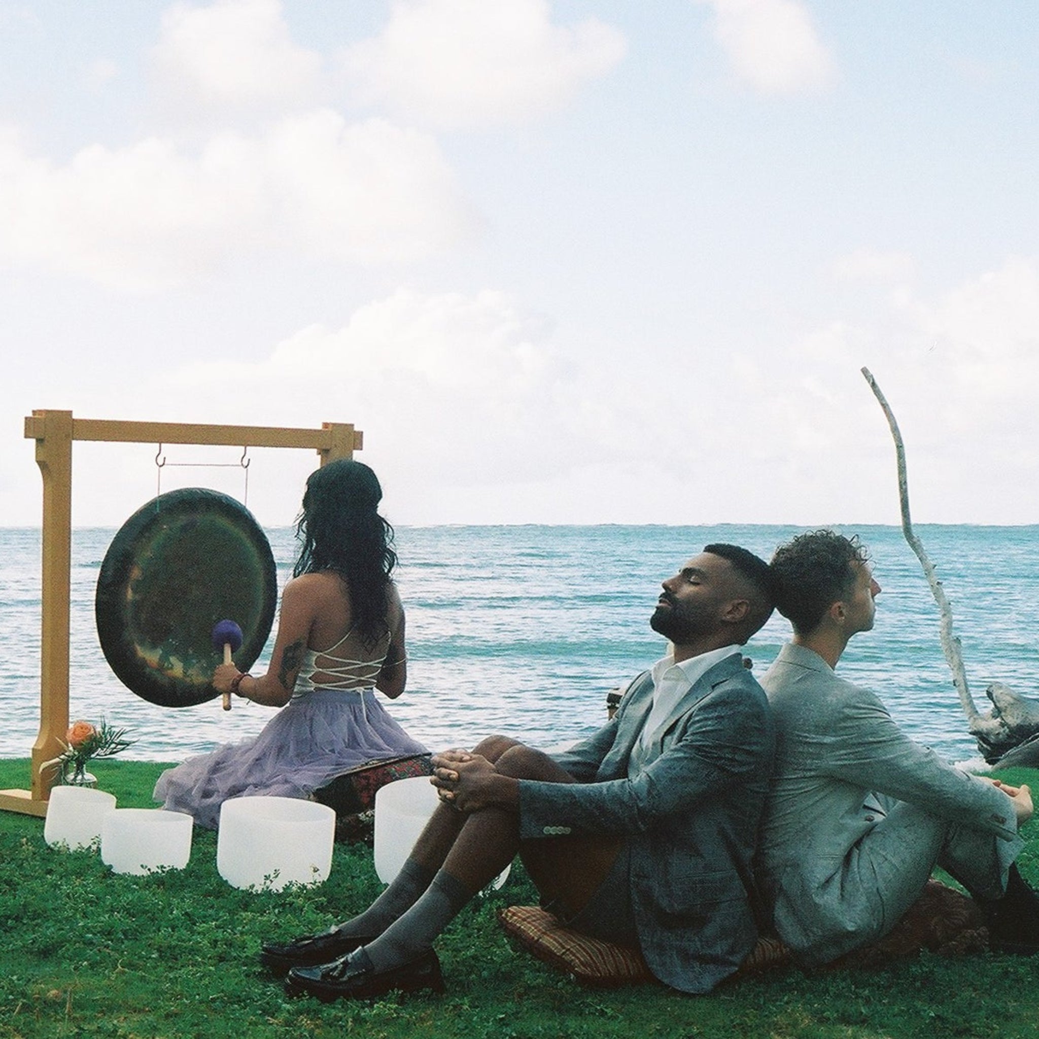 A group of people sitting on the grass by the ocean, playing musical instruments including a large gong, white drums, and a bow, under a cloudy sky.
