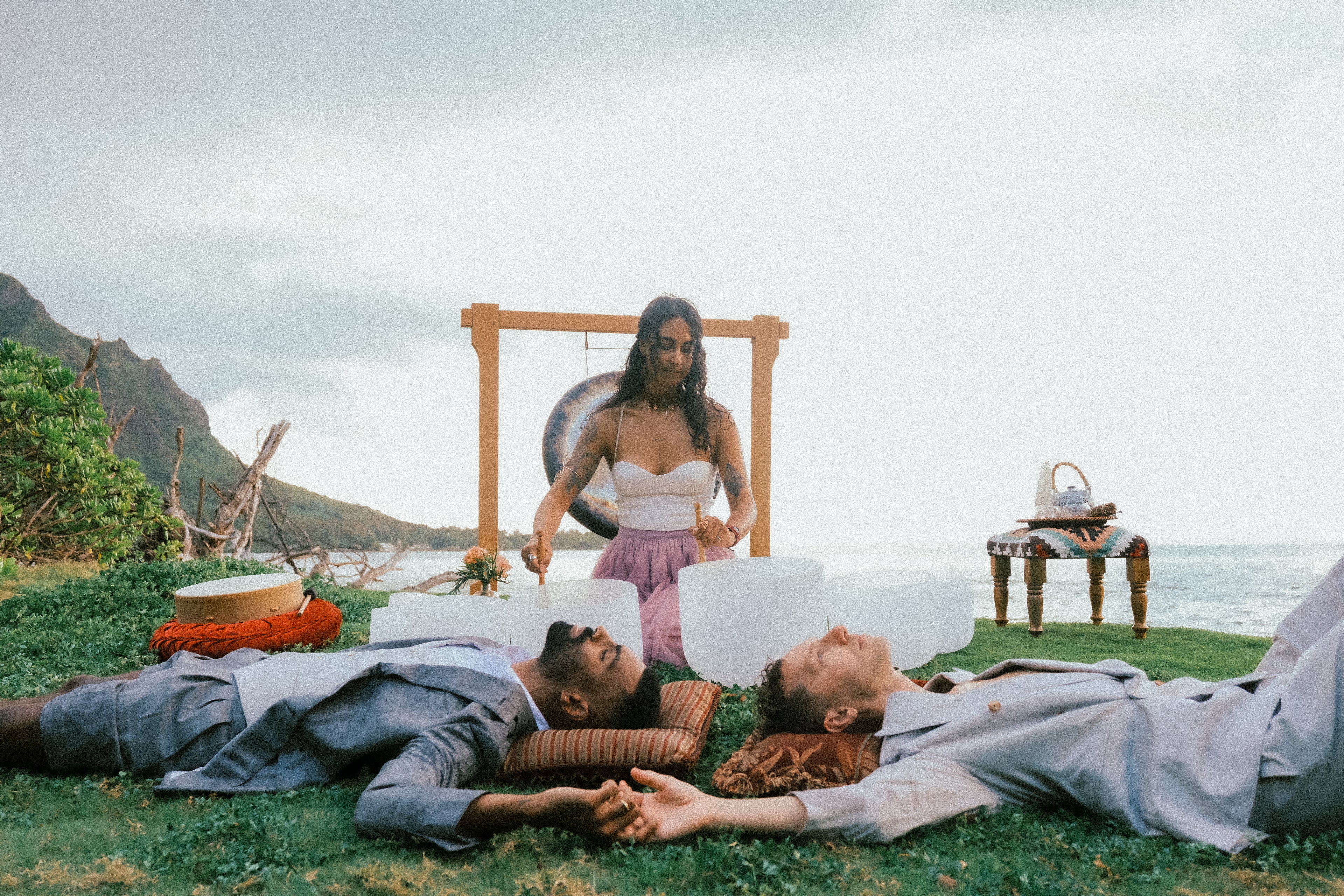 A woman stands behind two men lying on grass, holding white blocks, with a wooden gong frame and ocean in the background. The scene is outdoors on a beach or grassy coastal area with mountains and sky visible.