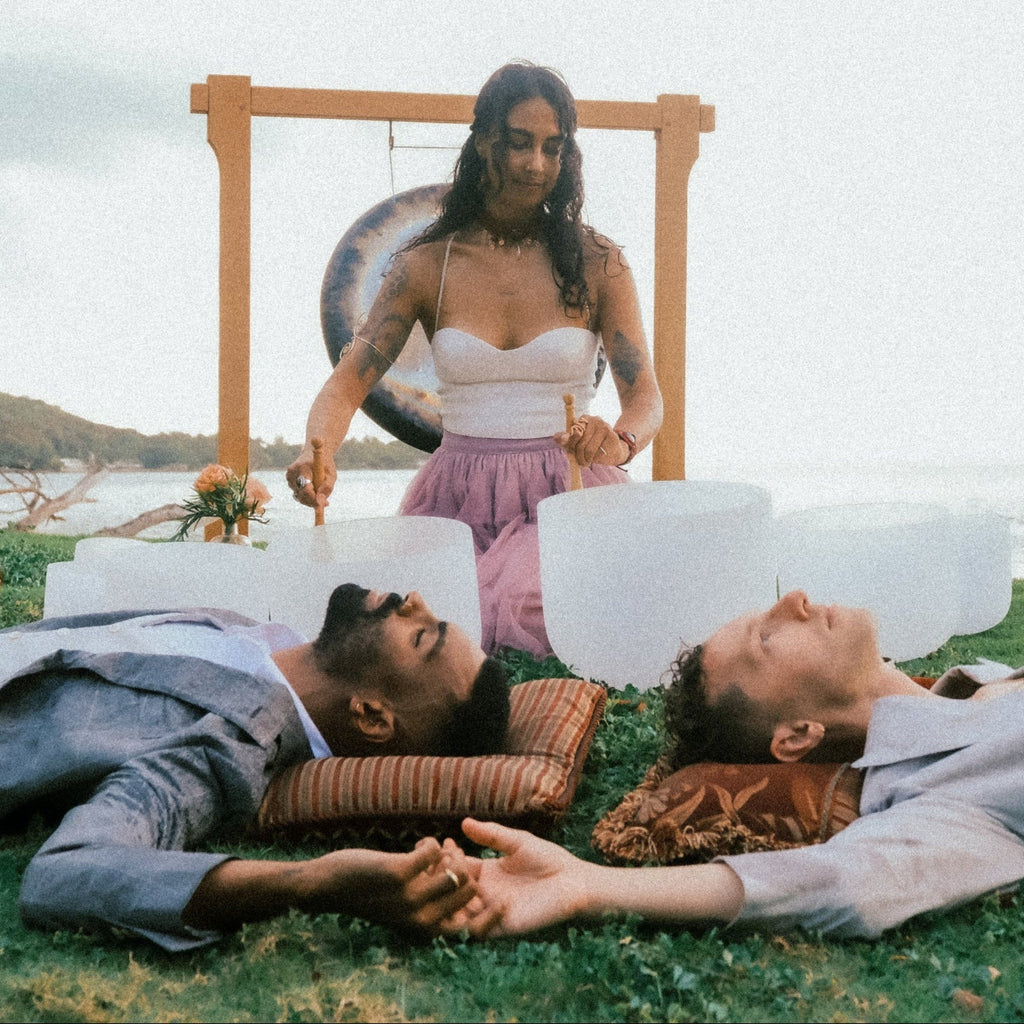 Two people lying on the grass holding hands with a woman standing behind them, all near a body of water.