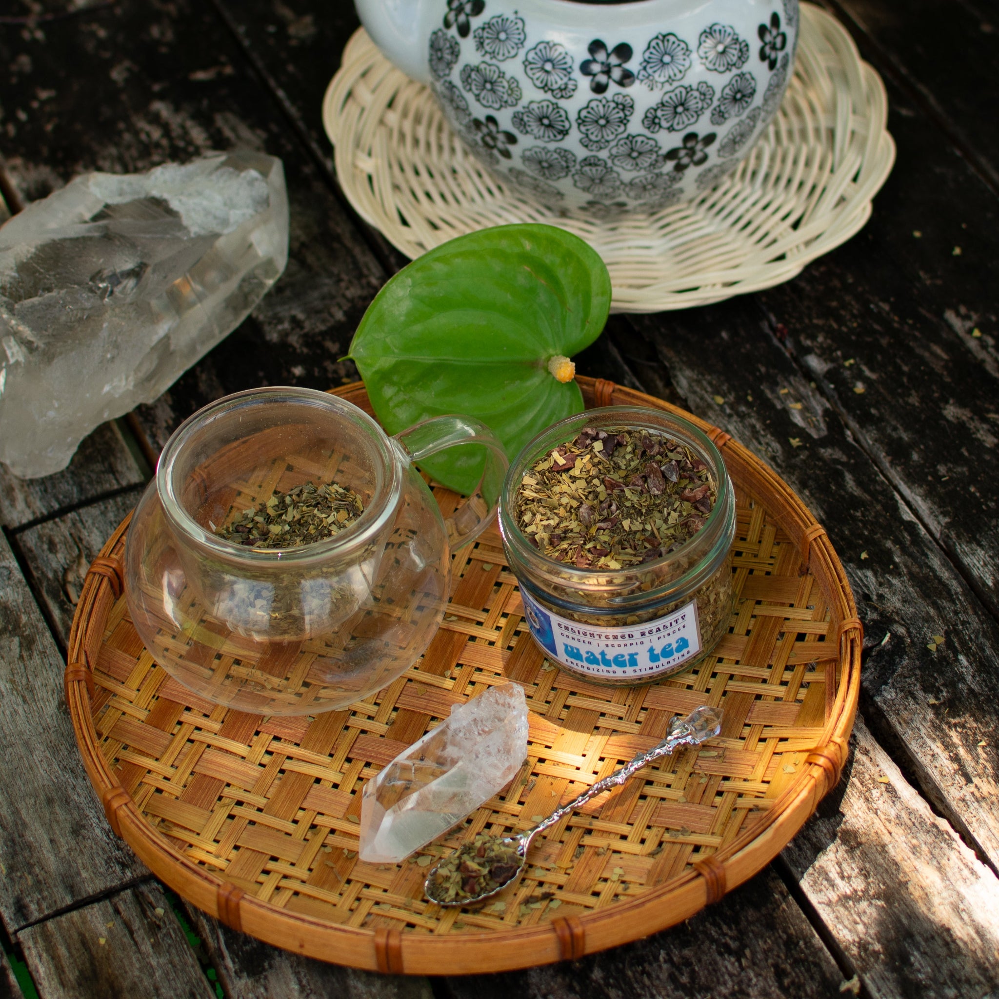 Glass teacup with loose herbal tea, crystal, and teapot on wooden table – Water Tea blend for energizing rituals